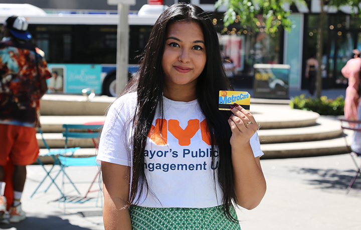 A person wearing a PEU t-shirt smiles at the camera and holds up a MetroCard
                                           